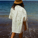 Image of woman on beach from behind, wearing the 'Seasons of Change' by Mary Ball Shirt in Green. The back of the shirt features green botanical drawings. 