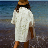 Image of woman on beach from behind, wearing the 'Seasons of Change' by Mary Ball Shirt in Green. The back of the shirt features green botanical drawings. 