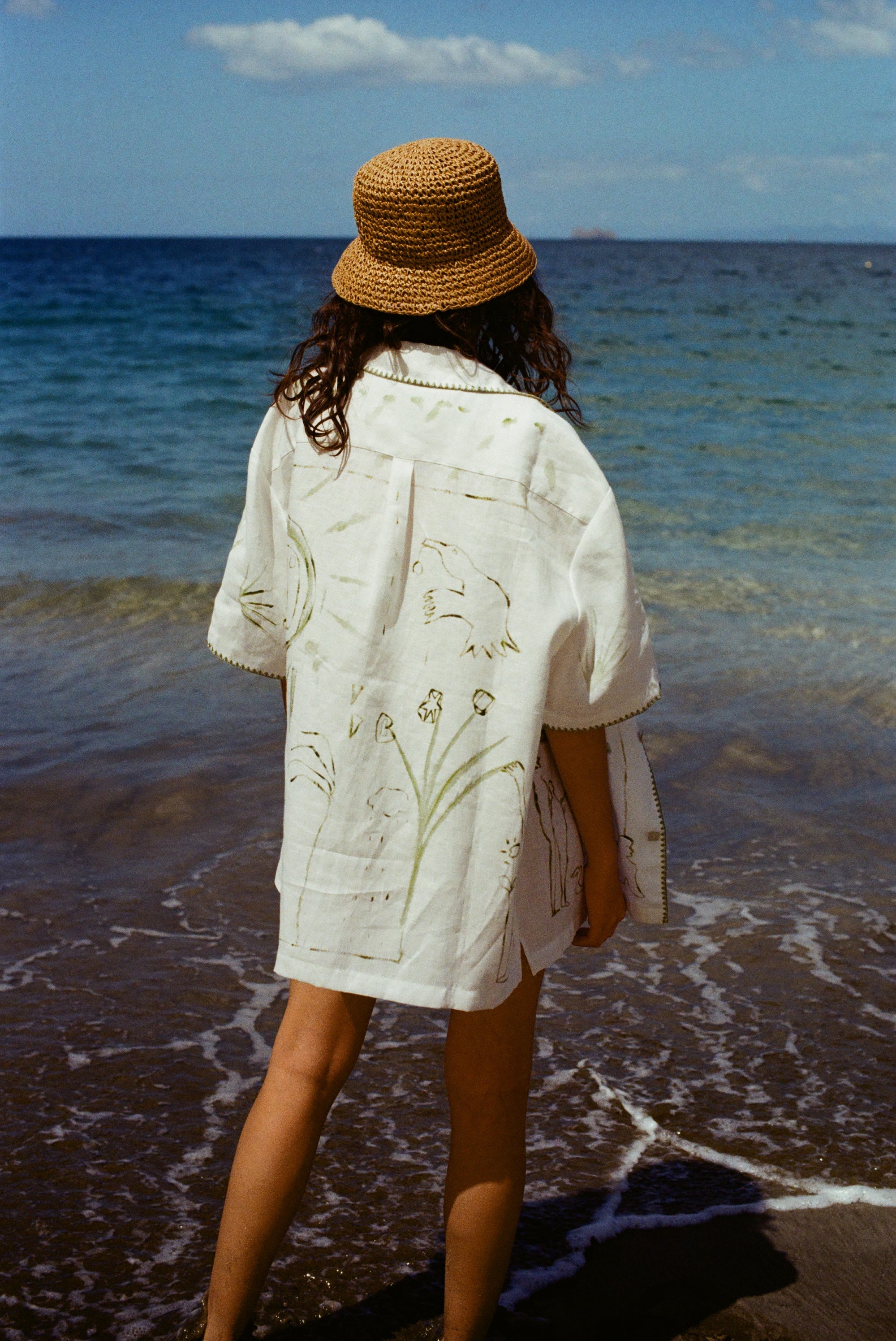 Image of woman on beach from behind, wearing the 'Seasons of Change' by Mary Ball Shirt in Green. The back of the shirt features green botanical drawings. 