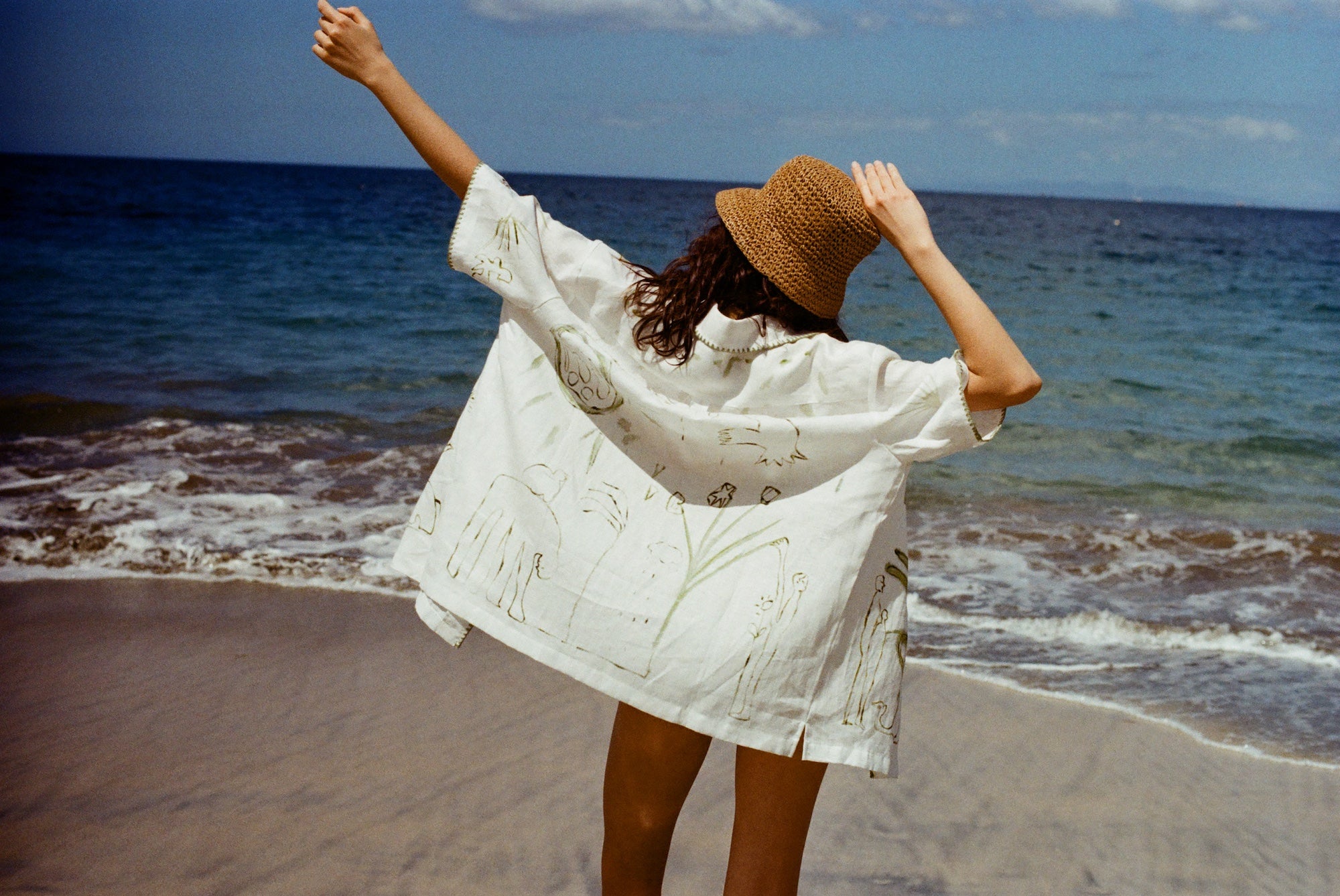Woman standing on beach facing the ocean with her back to us, while wearing the 'Seasons of Change' by Mary Ball Shirt in Green. Her arms are up in the air. 