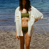 Women at the beach looks at the camera with a hand on her hip, wearing a green bikini, a white Tombolo button down shirt and a beige raffia hat 