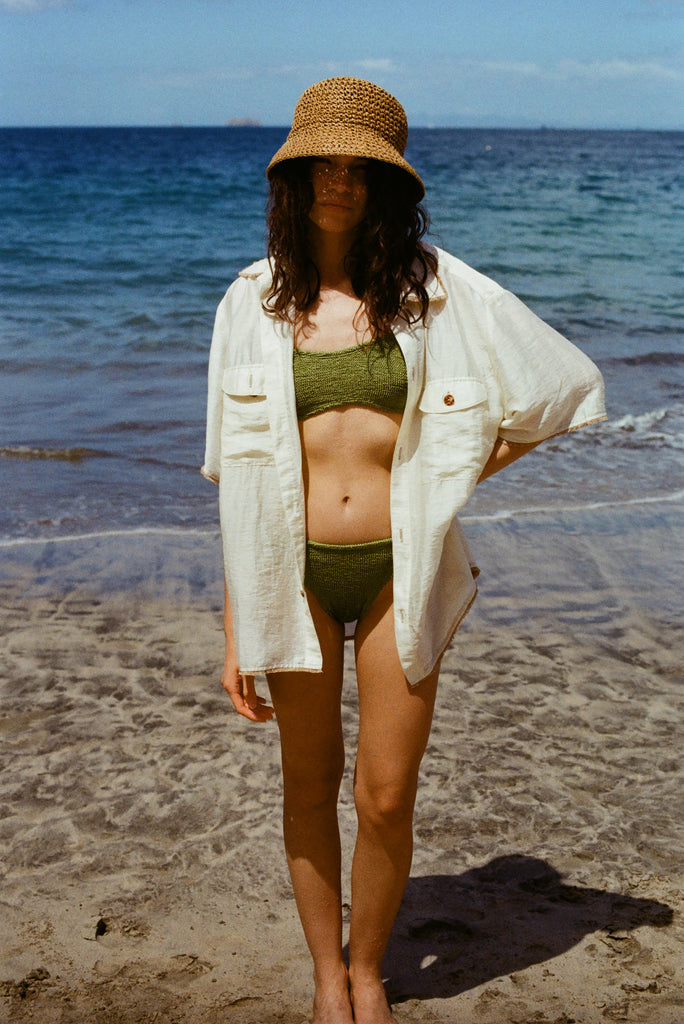 Women at the beach looks at the camera with a hand on her hip, wearing a green bikini, a white Tombolo button down shirt and a beige raffia hat 