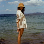 Woman at beach standing on rock, wearing beige raffia hat and white button down shirt