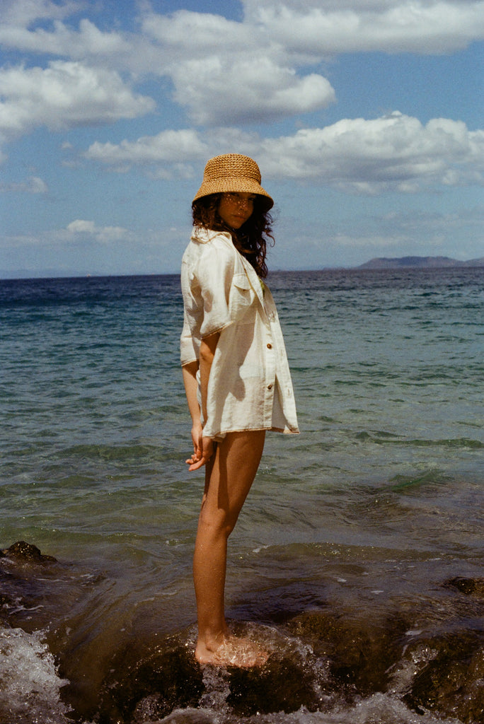 Woman at beach standing on rock, wearing beige raffia hat and white button down shirt