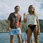 A woman stands at the front of a small boat wearing the 'Starfish Strut' Hand Towel Top—a mesh cream-colored shirt featuring a large starfish applique—paired with black swim bottoms. Next to her stands a man in a brown short-sleeve shirt and light blue printed swim trunks. In the background, another woman sits cross-legged in a light blue shirt with sand dollar details.