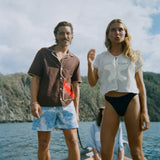 A woman stands at the front of a small boat wearing the 'Starfish Strut' Hand Towel Top—a mesh cream-colored shirt featuring a large starfish applique—paired with black swim bottoms. Next to her stands a man in a brown short-sleeve shirt and light blue printed swim trunks. In the background, another woman sits cross-legged in a light blue shirt with sand dollar details.