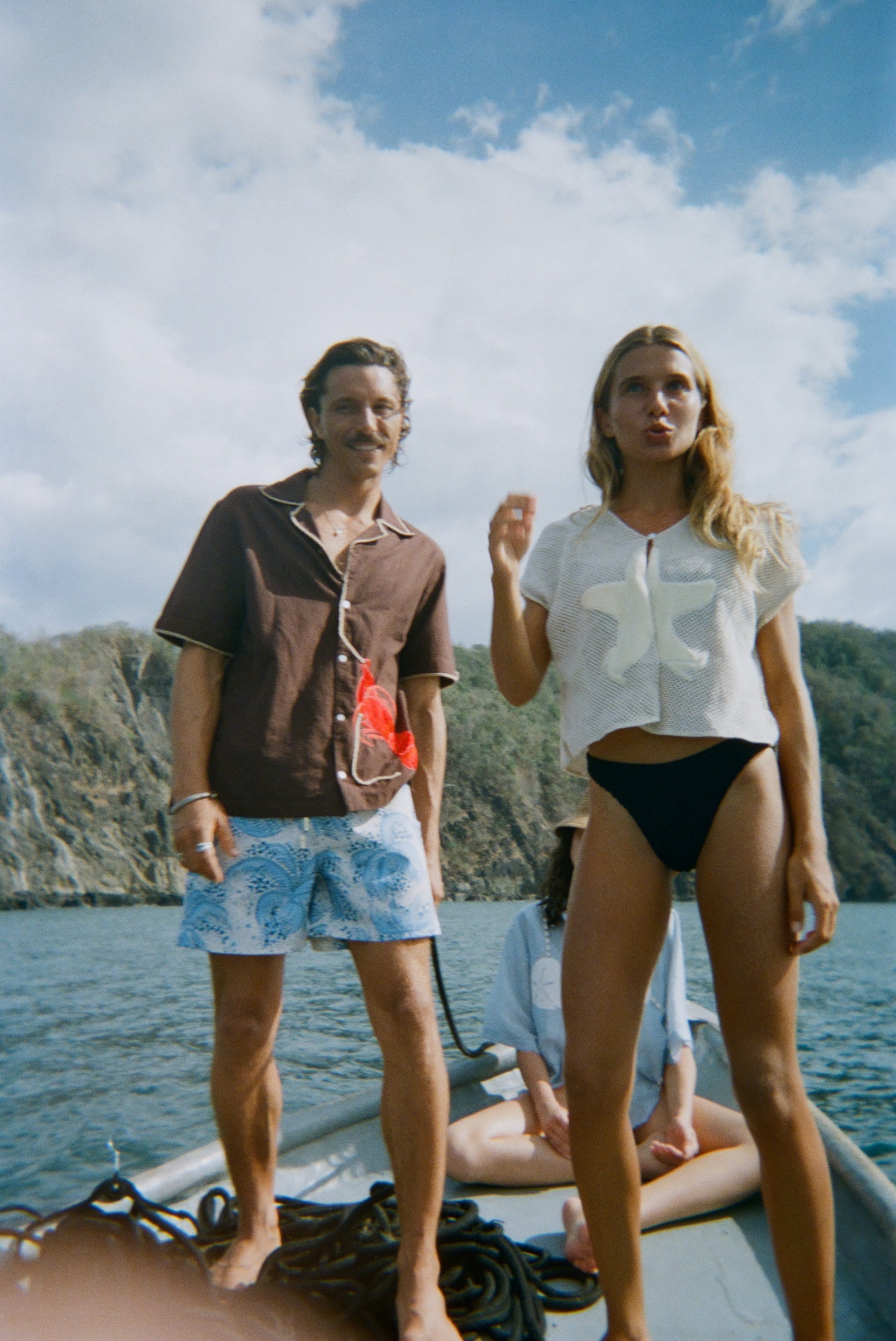 A woman stands at the front of a small boat wearing the 'Starfish Strut' Hand Towel Top—a mesh cream-colored shirt featuring a large starfish applique—paired with black swim bottoms. Next to her stands a man in a brown short-sleeve shirt and light blue printed swim trunks. In the background, another woman sits cross-legged in a light blue shirt with sand dollar details.