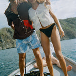 Two models stand side by side on the bow of a boat. The man wears a short-sleeve brown shirt with tan piping and a large red lobster applique on the lower half ("Bisque Averse") paired with blue paisley swim trunks. The woman wears a sheer white mesh top with a large cream starfish applique at the center ("Starfish Strut" Hand Towel Top) and black high-waisted bikini bottoms.