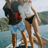 Two models stand side by side on the bow of a boat. The man wears a short-sleeve brown shirt with tan piping and a large red lobster applique on the lower half ("Bisque Averse") paired with blue paisley swim trunks. The woman wears a sheer white mesh top with a large cream starfish applique at the center ("Starfish Strut" Hand Towel Top) and black high-waisted bikini bottoms.