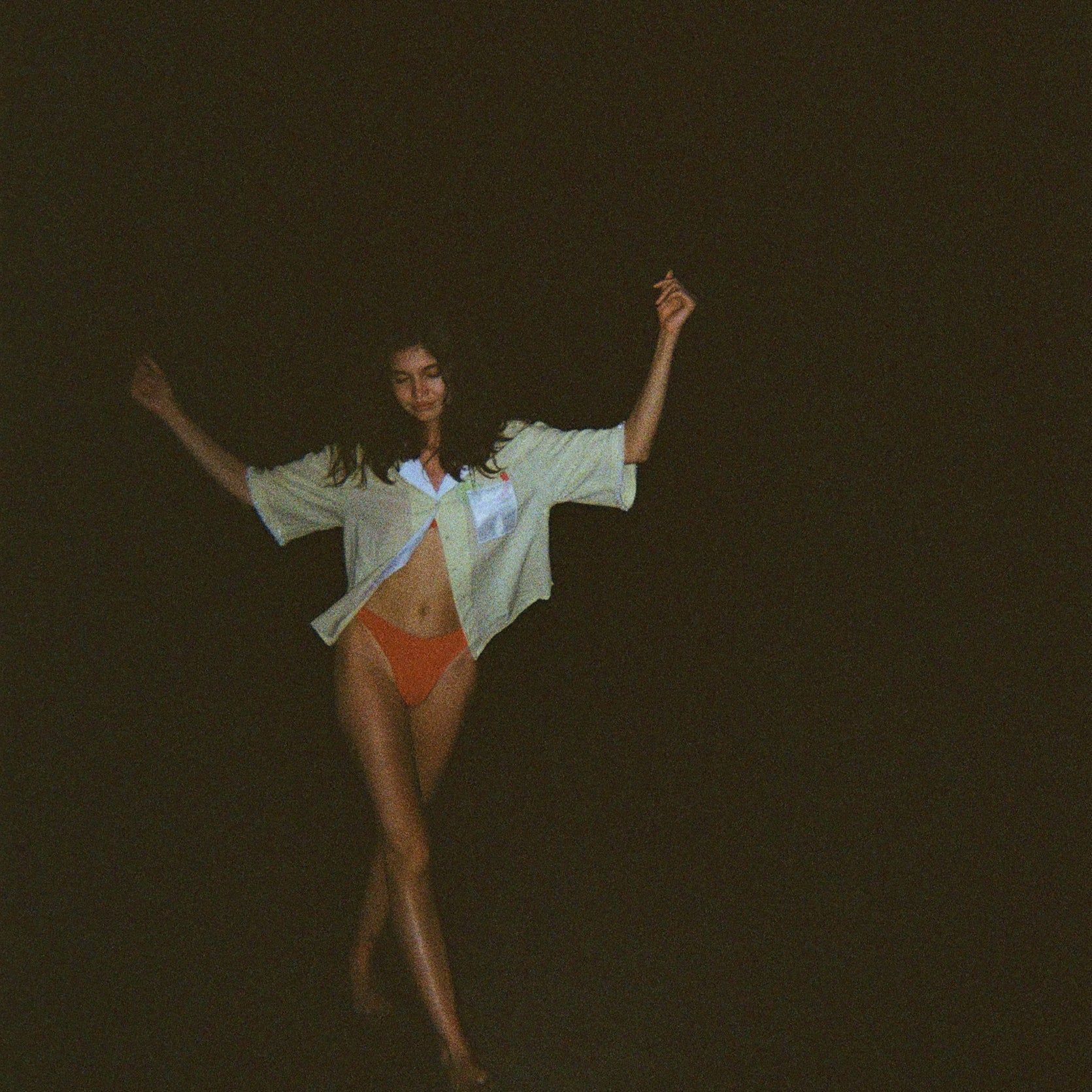 Woman dancing in the dark while wearing an open light yellow short-sleeve shirt with a white collar and pocket, over orange bikini bottoms.