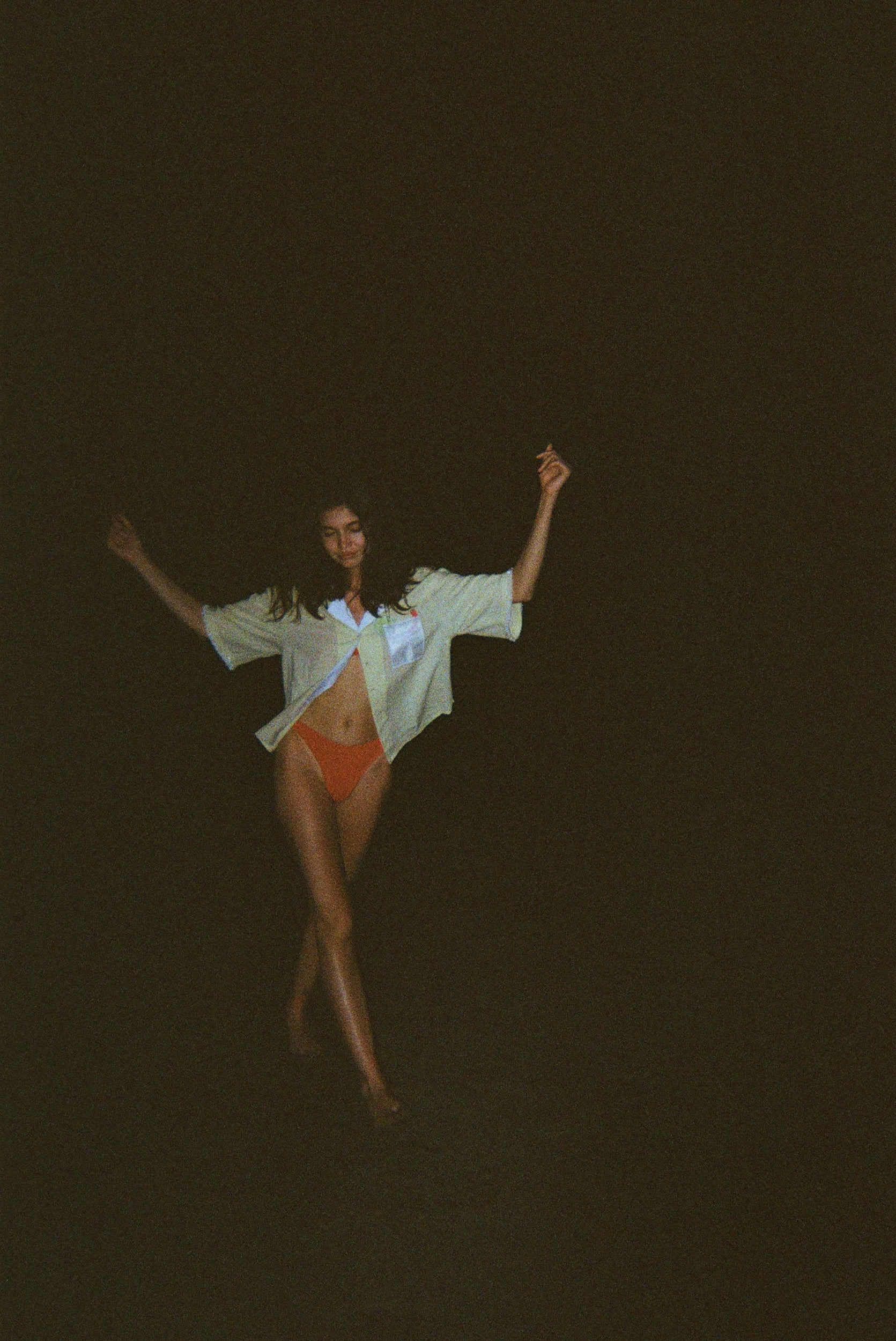 Woman dancing in the dark while wearing an open light yellow short-sleeve shirt with a white collar and pocket, over orange bikini bottoms.