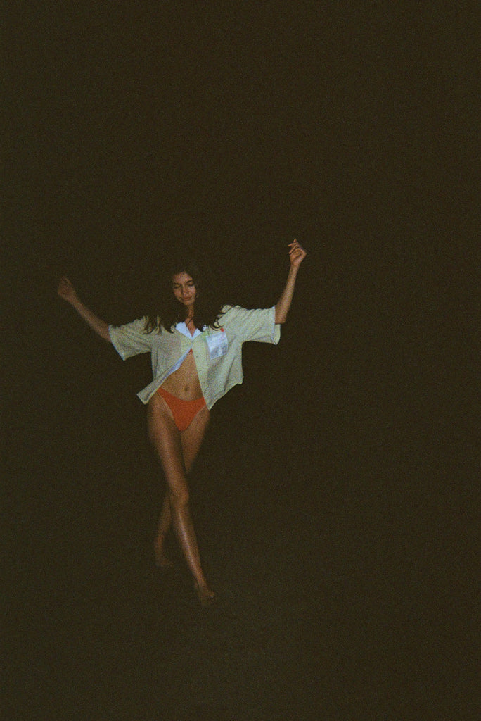 Woman dancing in the dark while wearing an open light yellow short-sleeve shirt with a white collar and pocket, over orange bikini bottoms.