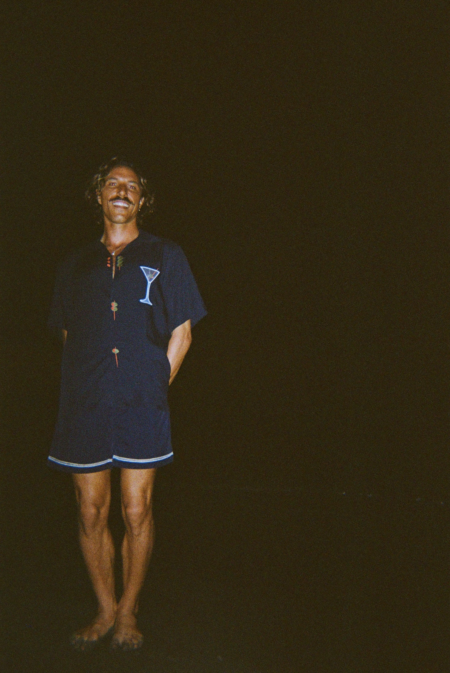 Model smiling widely, standing on the beach at night wearing the 'Extra Dirty' ~ Relaxed Fit Shirt and Navy swim trunks. 