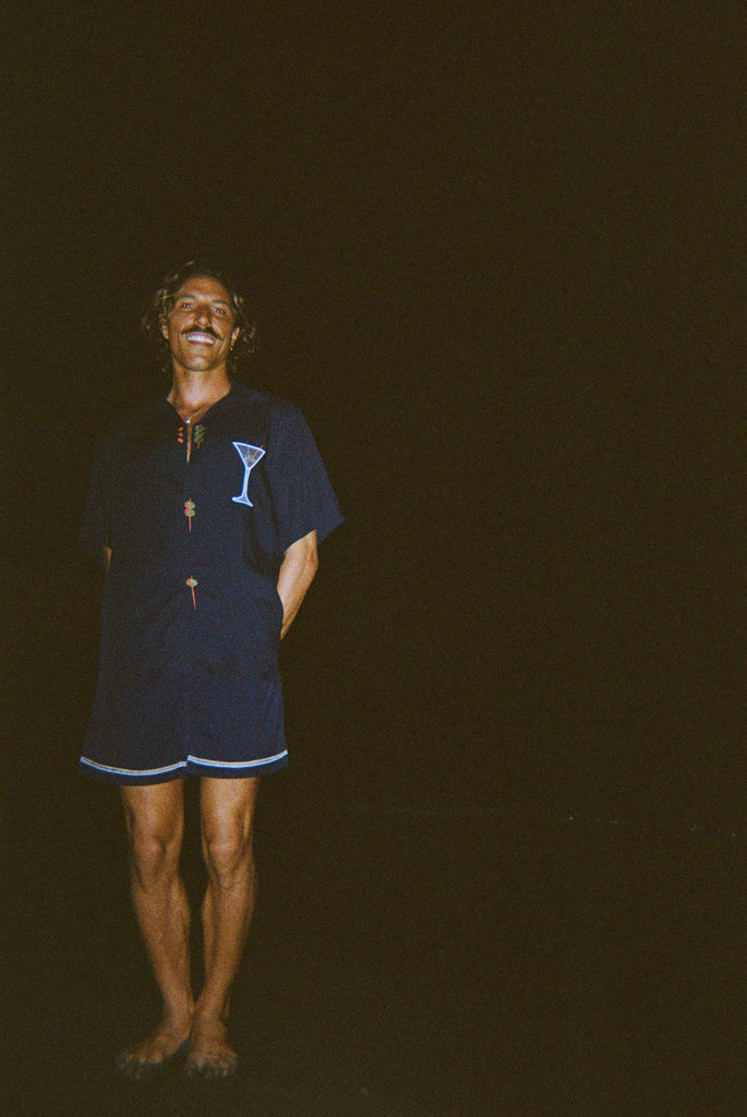 Model smiling widely, standing on the beach at night wearing the 'Extra Dirty' ~ Relaxed Fit Shirt and Navy swim trunks. 