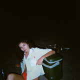 Woman sitting on dark beach while wearing an open light yellow short-sleeve shirt with a white collar and pocket, over orange bikini bottoms.