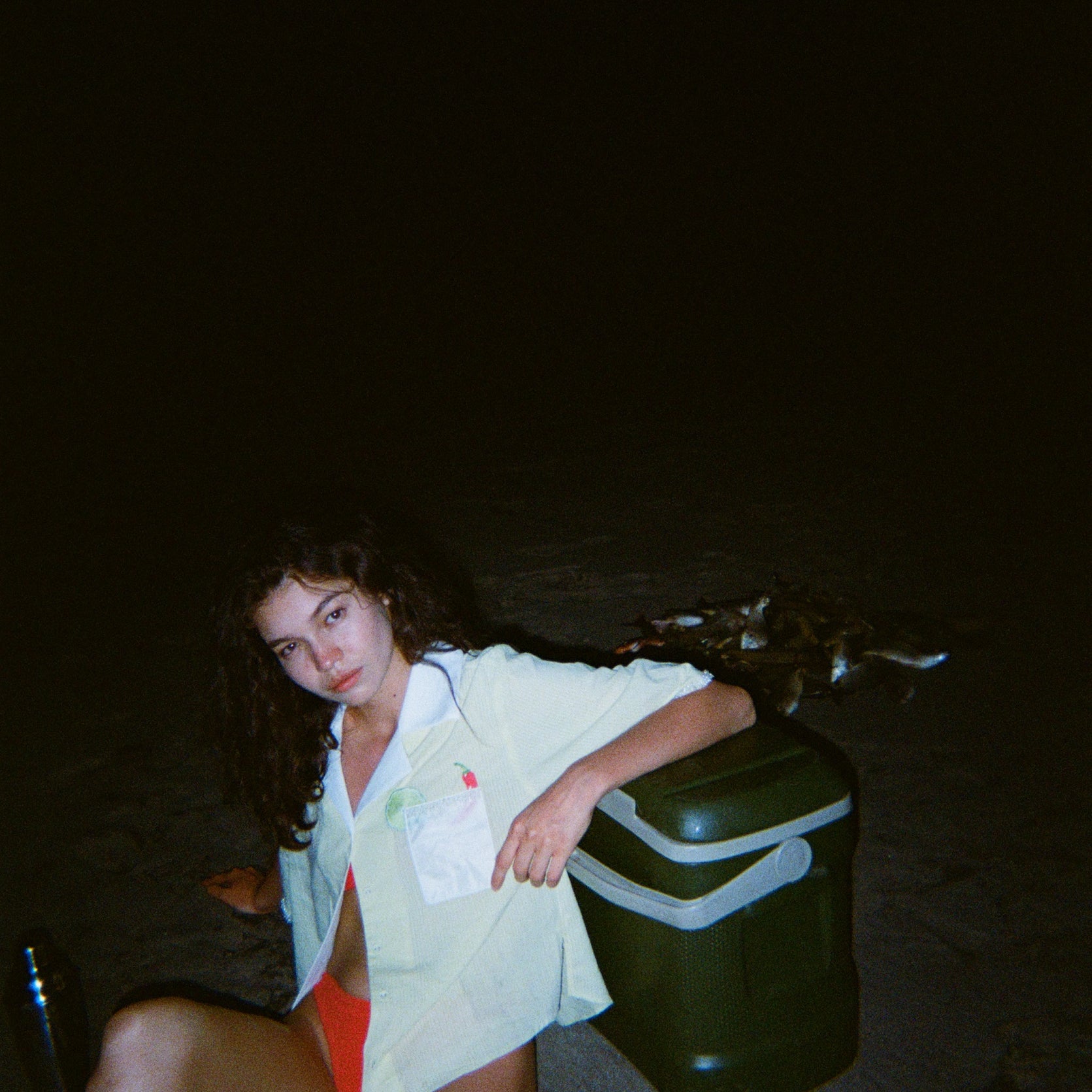 Woman sitting on dark beach while wearing an open light yellow short-sleeve shirt with a white collar and pocket, over orange bikini bottoms.