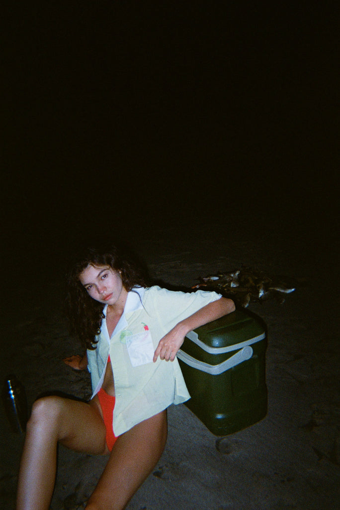 Woman sitting on dark beach while wearing an open light yellow short-sleeve shirt with a white collar and pocket, over orange bikini bottoms.