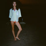 Woman standing on a dark beach while wearing an open light yellow short-sleeve shirt with a white collar and pocket, over orange bikini bottoms.