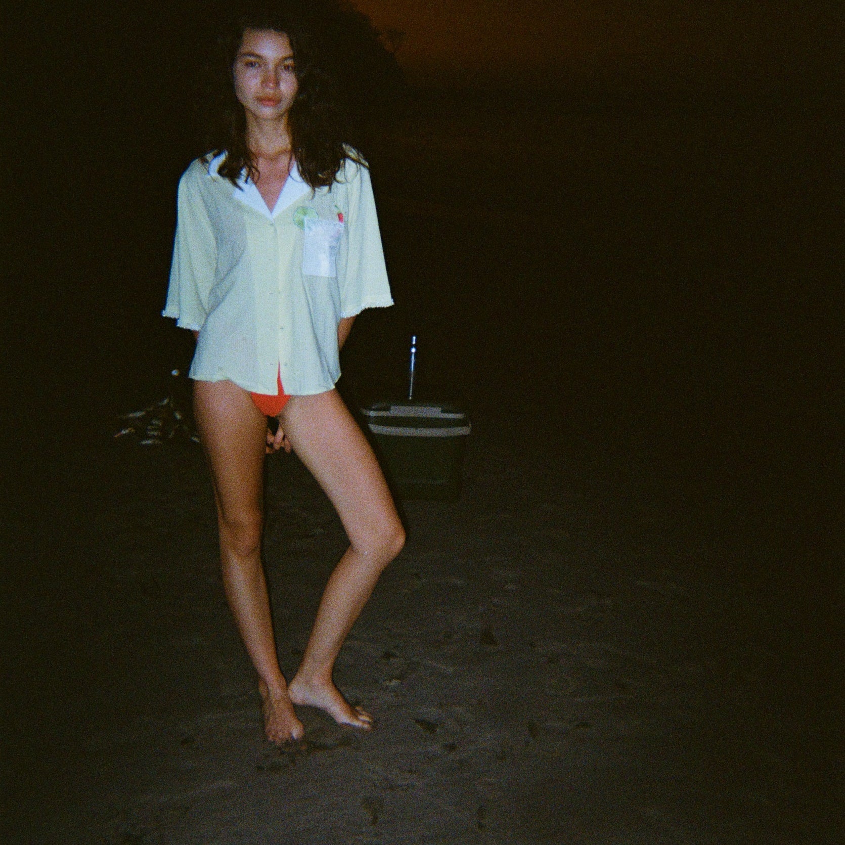 Woman standing on a dark beach while wearing an open light yellow short-sleeve shirt with a white collar and pocket, over orange bikini bottoms.