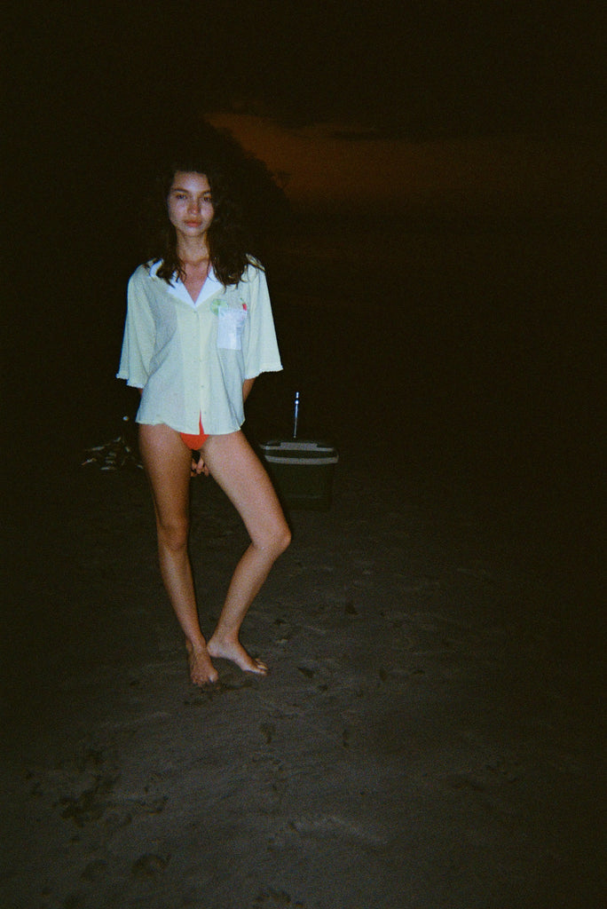 Woman standing on a dark beach while wearing an open light yellow short-sleeve shirt with a white collar and pocket, over orange bikini bottoms.