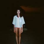 Woman standing on a dark beach while wearing an open light yellow short-sleeve shirt with a white collar and pocket, over orange bikini bottoms.