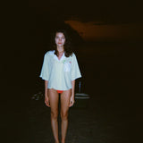 Woman standing on a dark beach while wearing an open light yellow short-sleeve shirt with a white collar and pocket, over orange bikini bottoms.