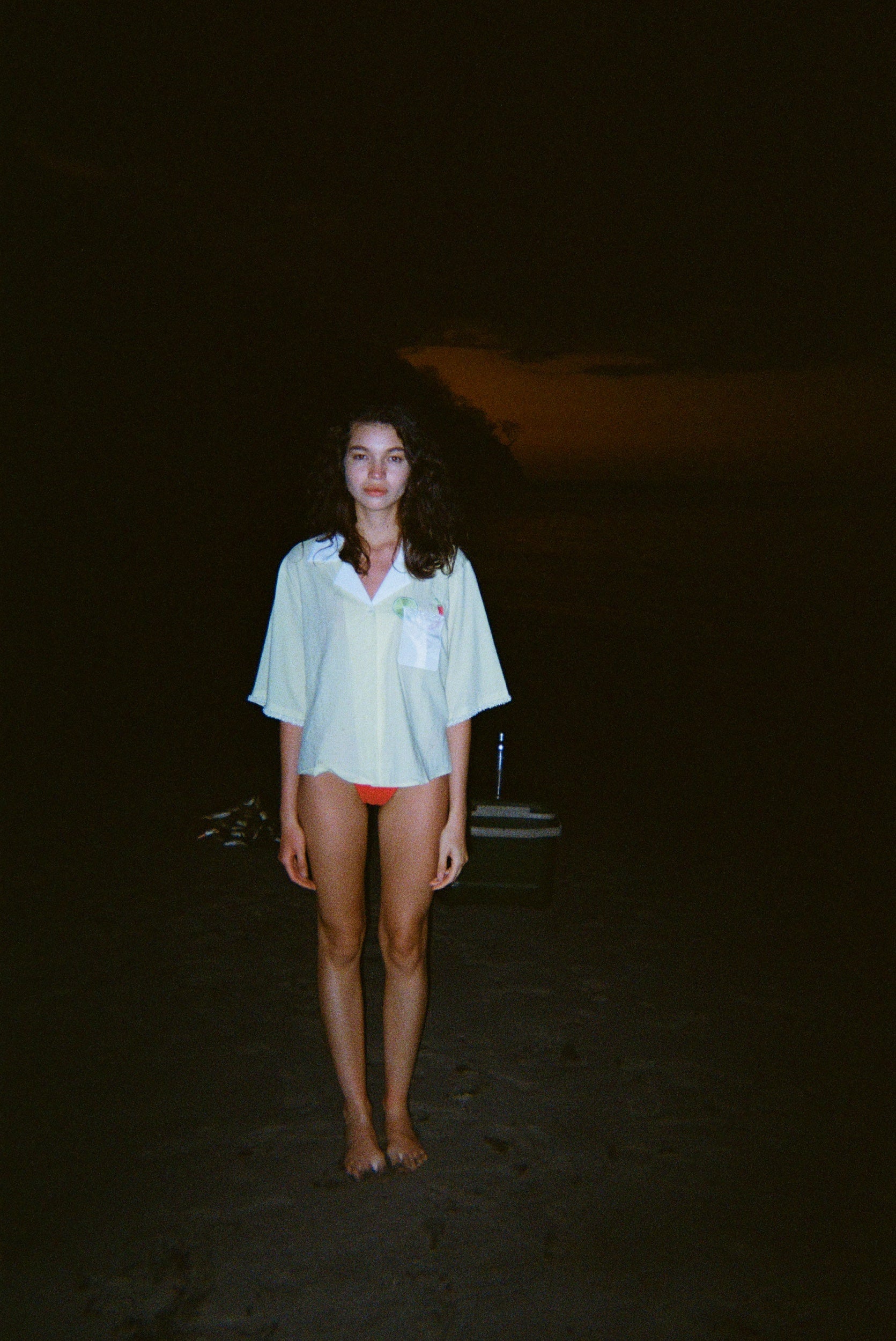 Woman standing on a dark beach while wearing an open light yellow short-sleeve shirt with a white collar and pocket, over orange bikini bottoms.