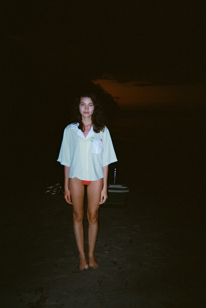 Woman standing on a dark beach while wearing an open light yellow short-sleeve shirt with a white collar and pocket, over orange bikini bottoms.
