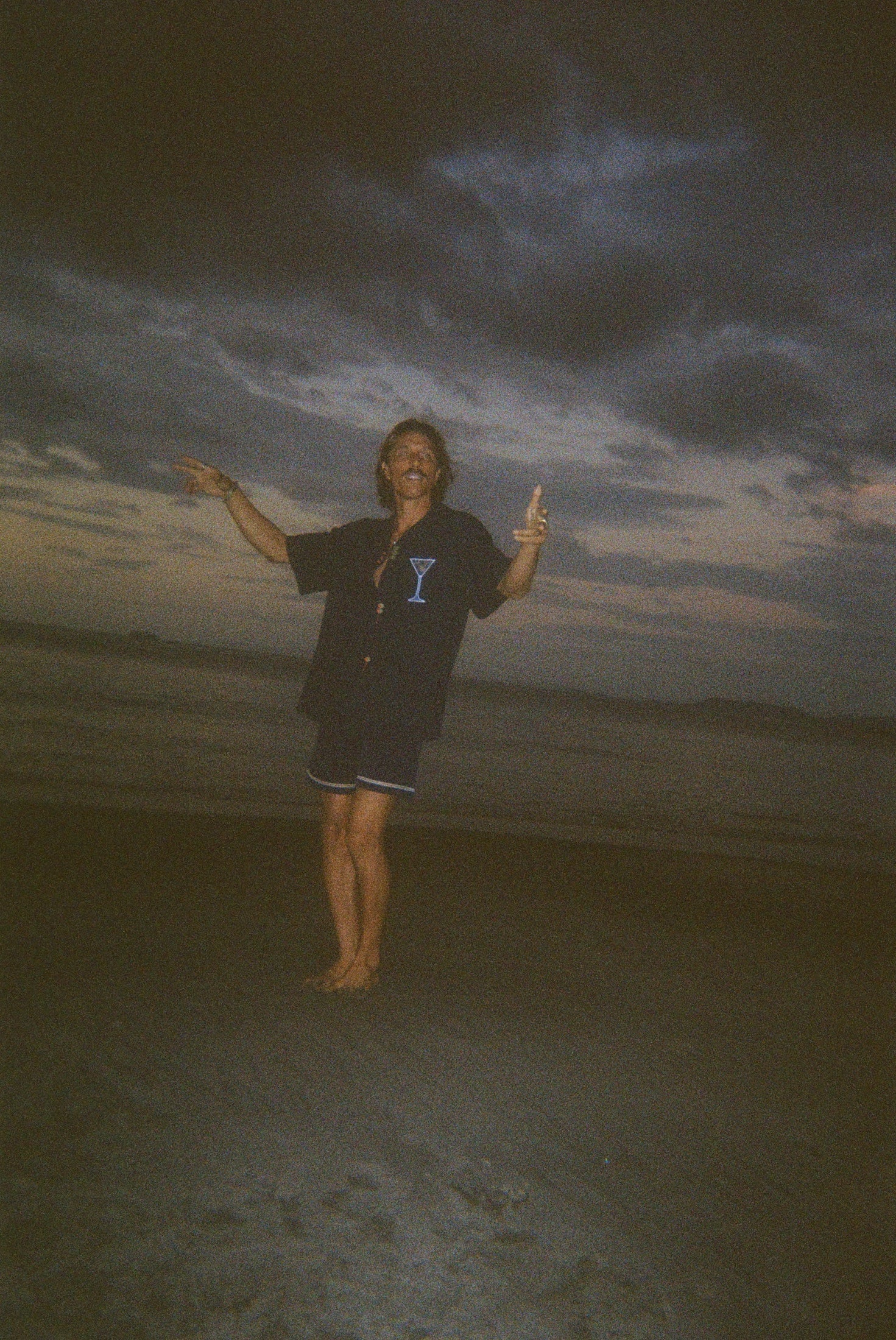 Male model dancing playfully on the beach, wearing the 'Extra Dirty' ~ Relaxed Fit Shirt—a navy short-sleeve button-up with embroidered martini and olive details—and navy swim trunks. The ocean and sand surround him.