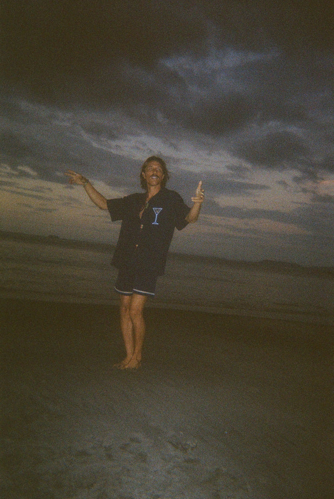 Male model dancing playfully on the beach, wearing the 'Extra Dirty' ~ Relaxed Fit Shirt—a navy short-sleeve button-up with embroidered martini and olive details—and navy swim trunks. The ocean and sand surround him.