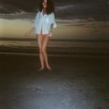 Woman dancing on a dark beach while wearing an open light yellow short-sleeve shirt with a white collar and pocket, over orange bikini bottoms.