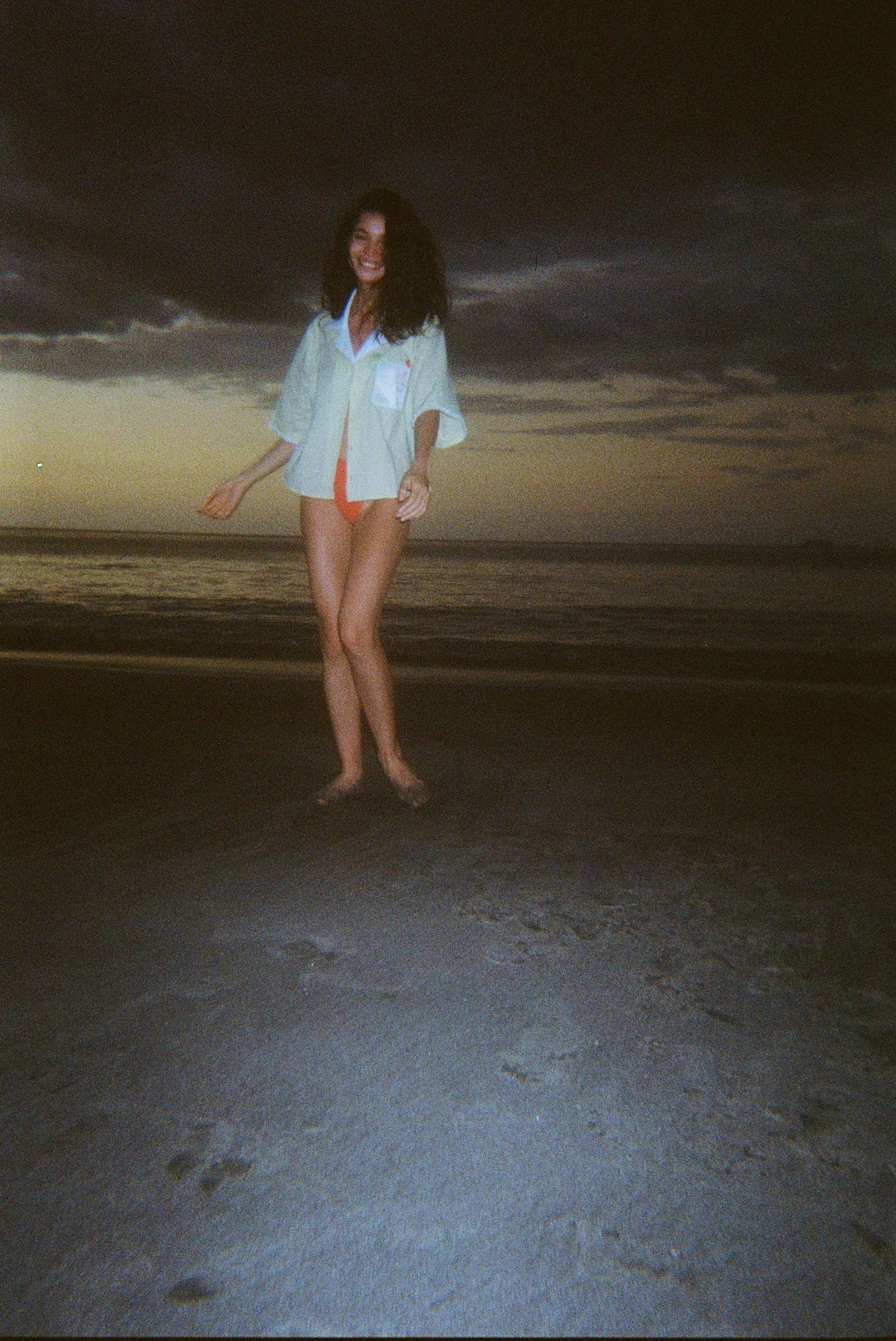 Woman dancing on a dark beach while wearing an open light yellow short-sleeve shirt with a white collar and pocket, over orange bikini bottoms.