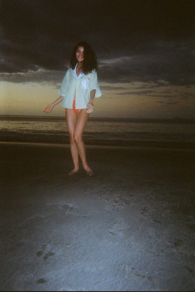 Woman dancing on a dark beach while wearing an open light yellow short-sleeve shirt with a white collar and pocket, over orange bikini bottoms.