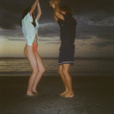 Man and woman dancing barefoot on the beach at dusk. The woman wears a light yellow shirt and orange bikini bottoms, and the man wears a navy outfit with shorts. 