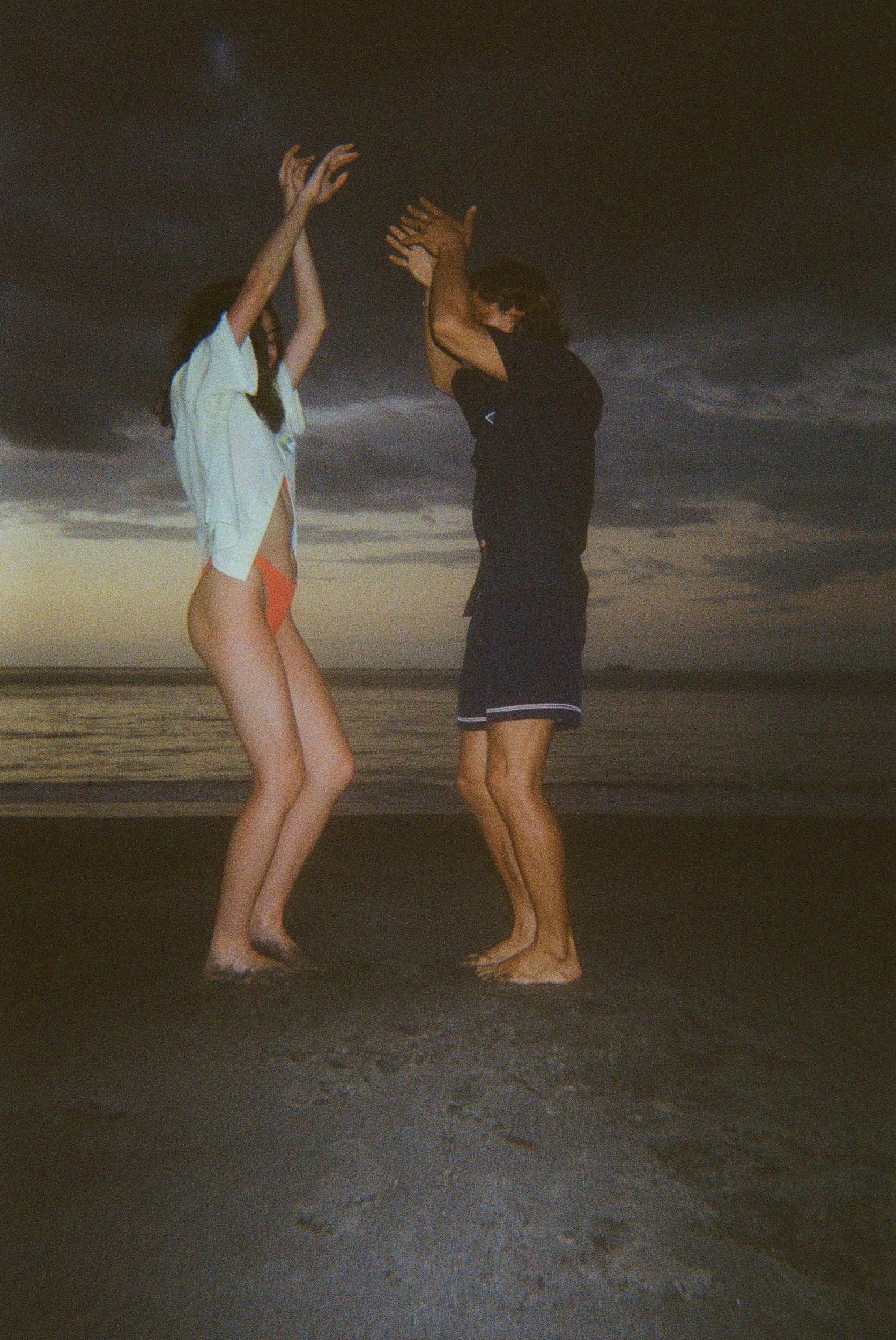 Man and woman dancing barefoot on the beach at dusk. The woman wears a light yellow shirt and orange bikini bottoms, and the man wears a navy outfit with shorts. 