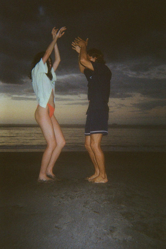 Man and woman dancing barefoot on the beach at dusk. The woman wears a light yellow shirt and orange bikini bottoms, and the man wears a navy outfit with shorts. 