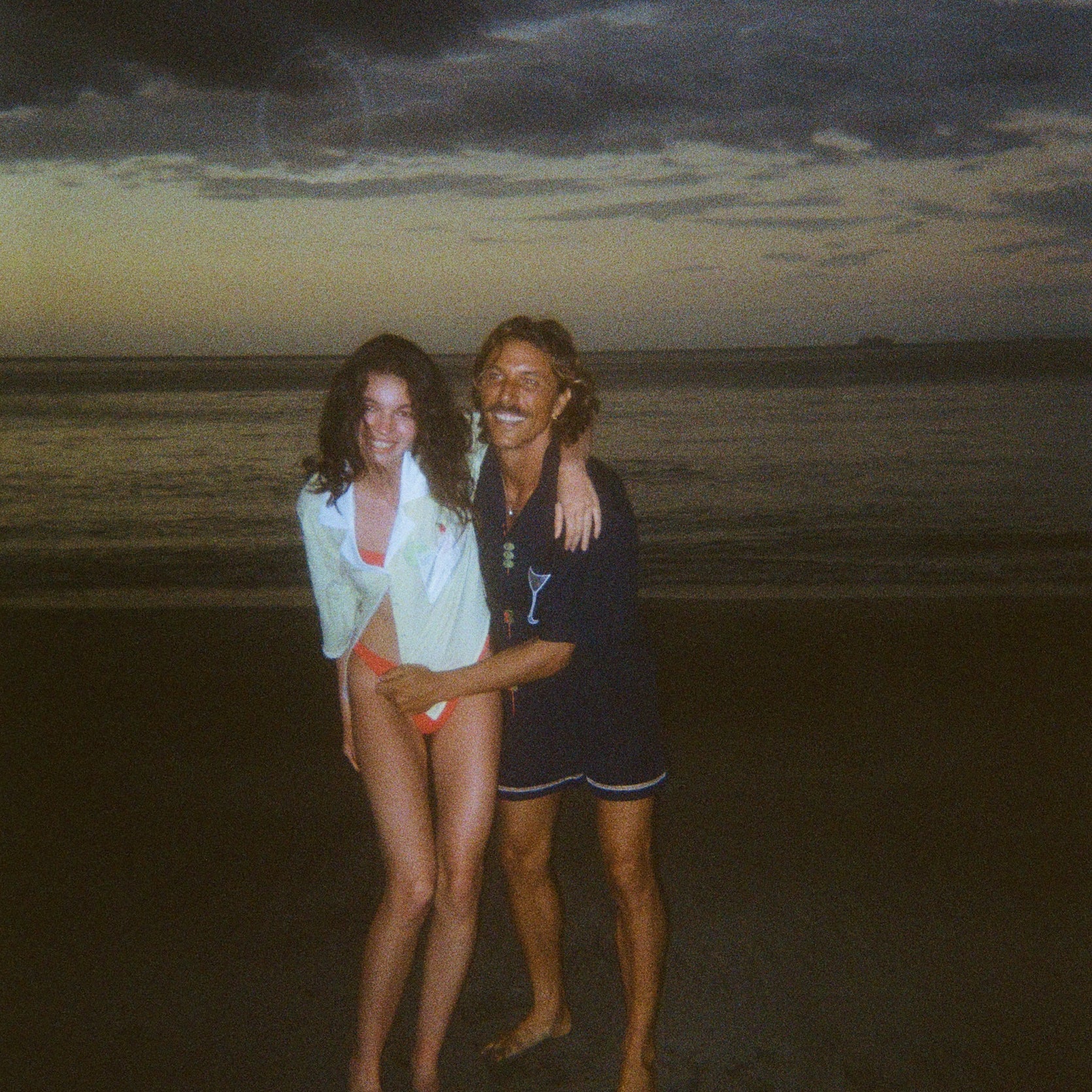Smiling man and woman standing on the beach at dusk. The woman wears an open light yellow shirt over orange bikini bottoms, and the man wears a navy shirt featuring a martini glass and navy swim trunks.