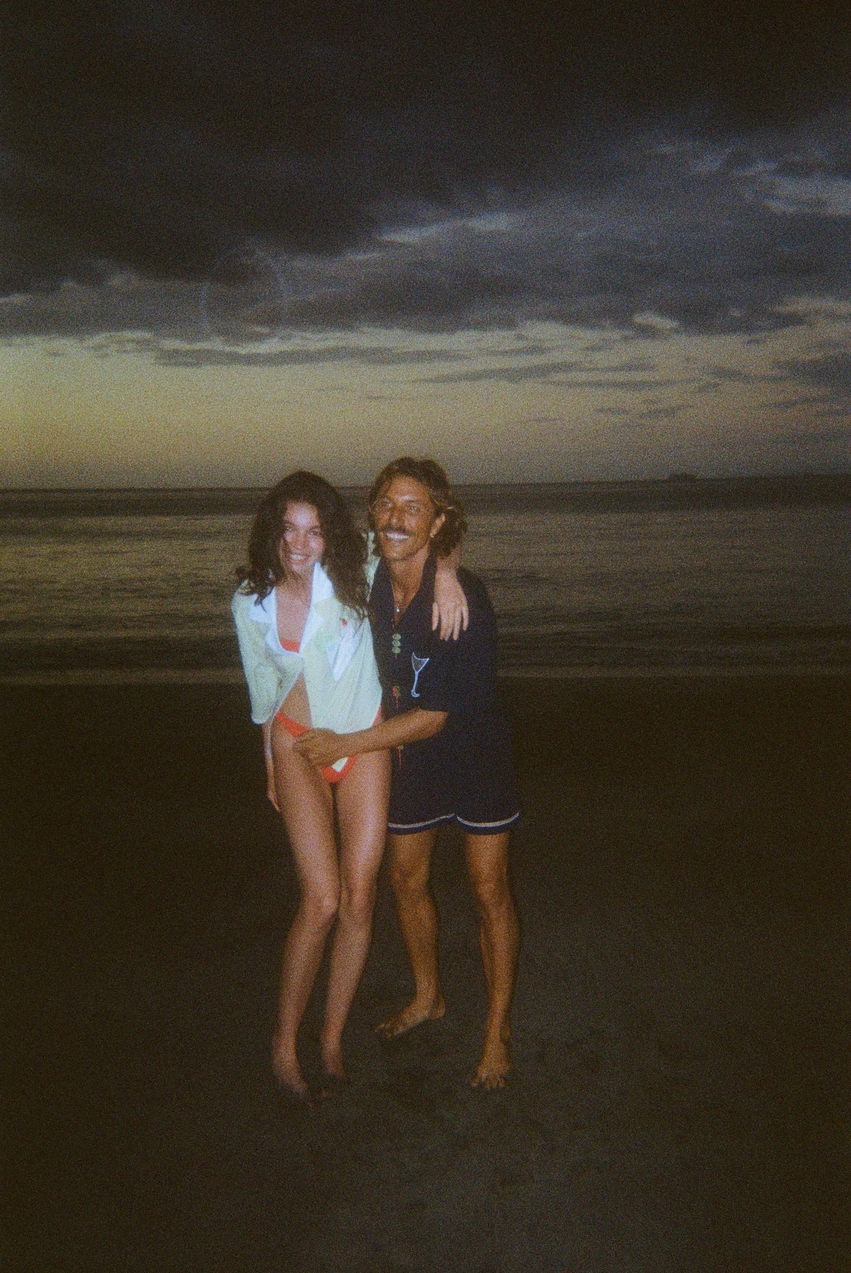 Smiling man and woman standing on the beach at dusk. The woman wears an open light yellow shirt over orange bikini bottoms, and the man wears a navy shirt featuring a martini glass and navy swim trunks.