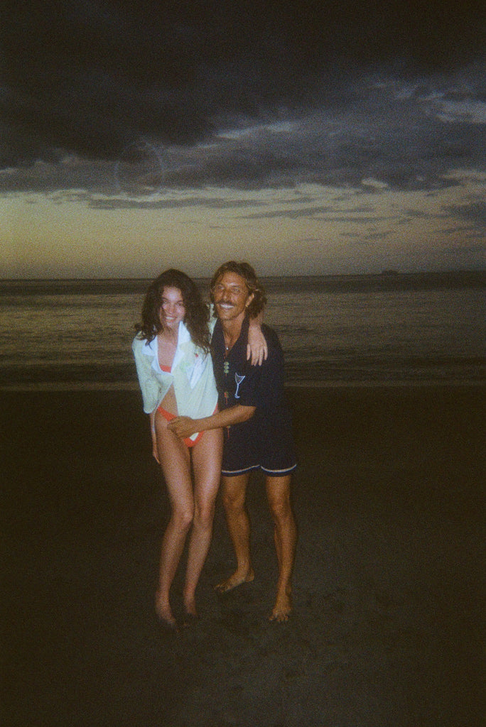 Smiling man and woman standing on the beach at dusk. The woman wears an open light yellow shirt over orange bikini bottoms, and the man wears a navy shirt featuring a martini glass and navy swim trunks.