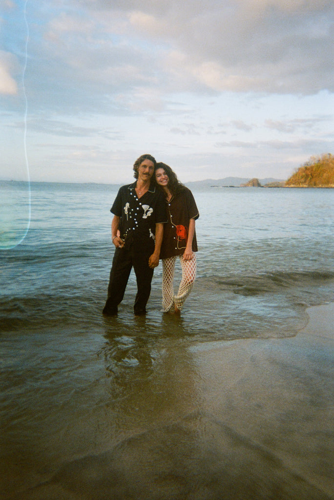 Two models stand ankle-deep in ocean water at sunset. The male model wears a black short-sleeve button-down shirt embroidered with two seahorses ('Tip your Bartenders' Cabana), paired with matching black trousers. The female model wears a brown short-sleeve shirt with beige piping and a large red lobster applique on the lower front ("Bisque Averse"), paired with white open-knit pants. They stand close together with the woman's hand resting gently on the man's shoulder.