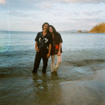 Two people standing ankle-deep in the ocean at sunset, smiling and leaning into each other. The man wears the black button down shirt and dark pants; the woman wears a dark shirt with a red lobster design and cream open-knit pants. Calm water surrounds them, with distant hills and a pastel sky in the background.