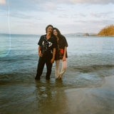 Two people standing ankle-deep in the ocean at sunset, smiling and leaning into each other. The man wears the black button down shirt and dark pants; the woman wears a dark shirt with a red lobster design and cream open-knit pants. Calm water surrounds them, with distant hills and a pastel sky in the background.