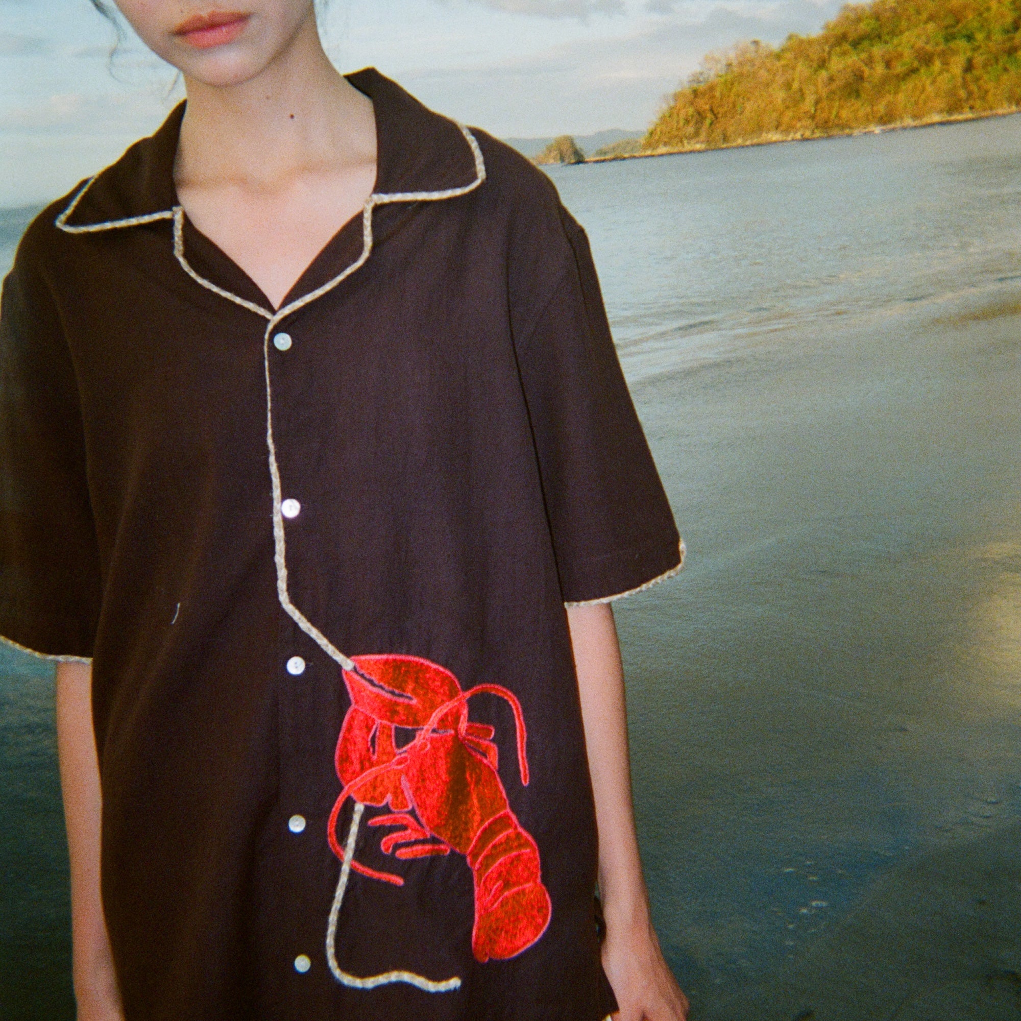 A woman stands on the beach at sunset wearing the "Bisque Averse" short-sleeve shirt—dark brown with contrast beige piping and a red textured lobster applique on the lower left side, connected by a rope-shaped trim that trails up the button placket. She pairs the shirt with white fishnet pants. 