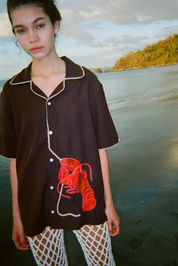 A woman stands on the beach at sunset wearing the "Bisque Averse" short-sleeve shirt—dark brown with contrast beige piping and a red textured lobster applique on the lower left side, connected by a rope-shaped trim that trails up the button placket. She pairs the shirt with white fishnet pants. 