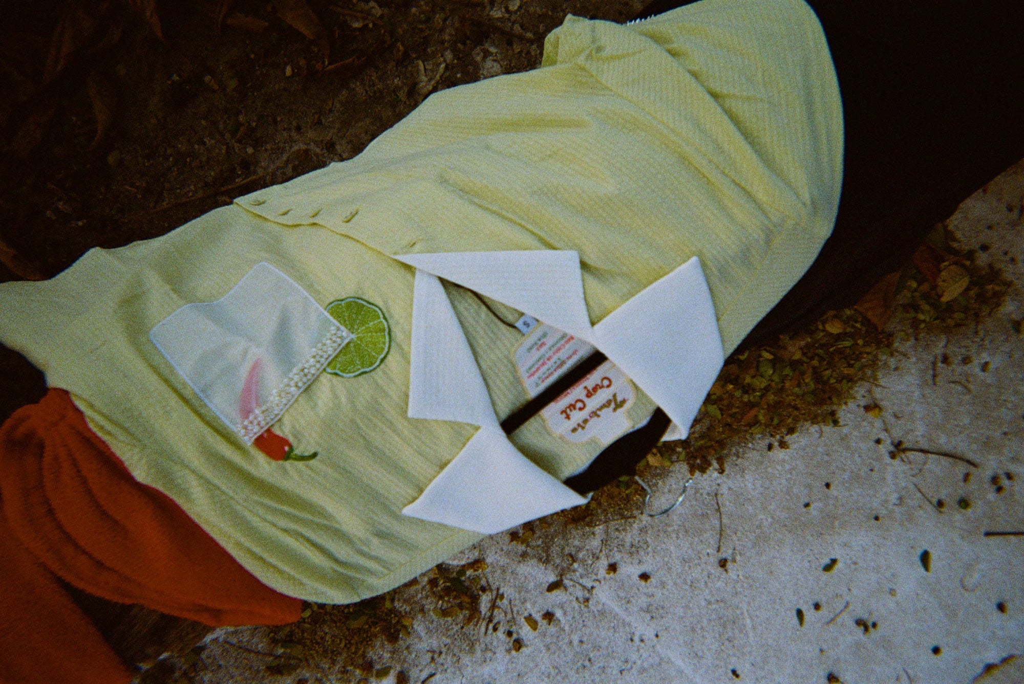 Light yellow short-sleeve shirt with a white collar laid flat on the ground. The shirt has an embroidered lime and red chili pepper near a white pocket with pearl trim. A red towel or garment is partially visible underneath.