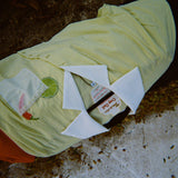 Light yellow short-sleeve shirt with a white collar laid flat on the ground. The shirt has an embroidered lime and red chili pepper near a white pocket with pearl trim. A red towel or garment is partially visible underneath.
