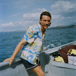 A man with wavy brown hair and a mustache leans against the edge of a boat, wearing a vibrant short-sleeve button-up shirt with a fishing-themed print featuring leaping fish and swirling ocean waves in blue, green, yellow, and red (Our 'Perfect Cast' Shirt). He pairs it with navy shorts that have white embroidered trim. 