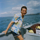 A man with wavy brown hair and a mustache leans against the edge of a boat, wearing a vibrant short-sleeve button-up shirt with a fishing-themed print featuring leaping fish and swirling ocean waves in blue, green, yellow, and red (Our 'Perfect Cast' Shirt). He pairs it with navy shorts that have white embroidered trim. 