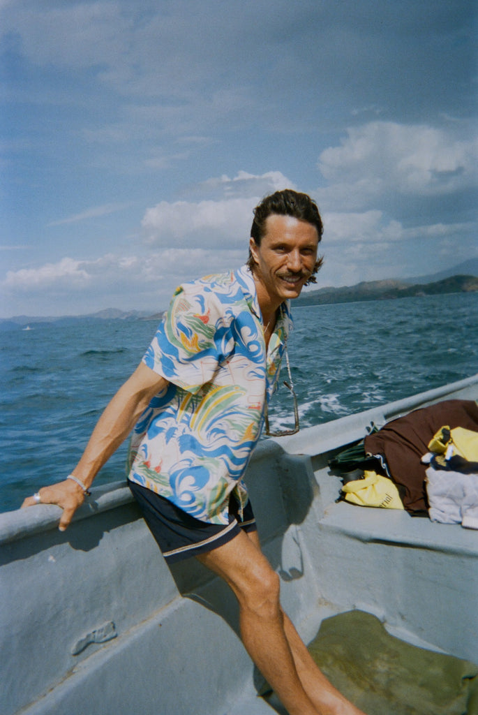 A man with wavy brown hair and a mustache leans against the edge of a boat, wearing a vibrant short-sleeve button-up shirt with a fishing-themed print featuring leaping fish and swirling ocean waves in blue, green, yellow, and red (Our 'Perfect Cast' Shirt). He pairs it with navy shorts that have white embroidered trim. 