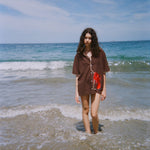 Female modeled pictured walking towards the camera at the beach, wearing the short-sleeved brown button-down ('Bisque Averse') over a swim suit. 